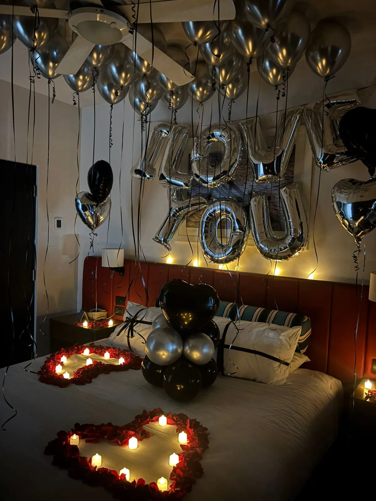 Hotel room decorated for a proposal return with silver I love you balloons, ceiling balloons and rose petal hearts lit by candles on the bed