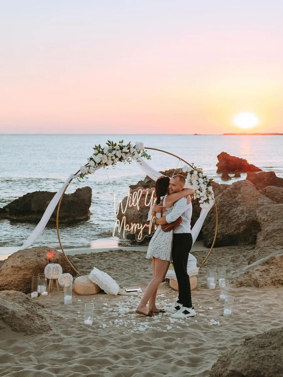 Newly engaged couple embracing under a circular floral arch on the beach at sunset
