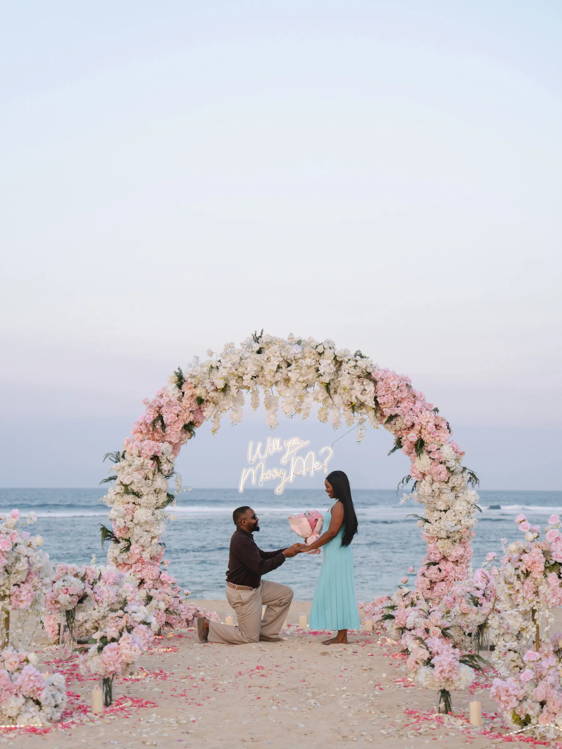 Pink and white circular floral arch with a neon Will You Marry Me sign on a Cabo beach