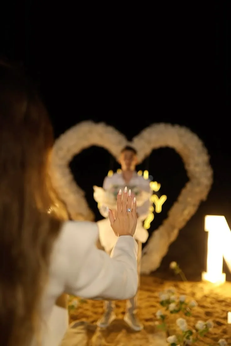 Engagement ring on a hand in front of an illuminated floral heart arch at night