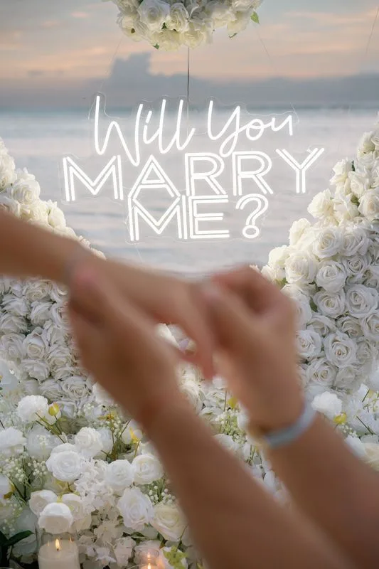 Hands with an engagement ring above white roses and a neon Will You Marry Me sign