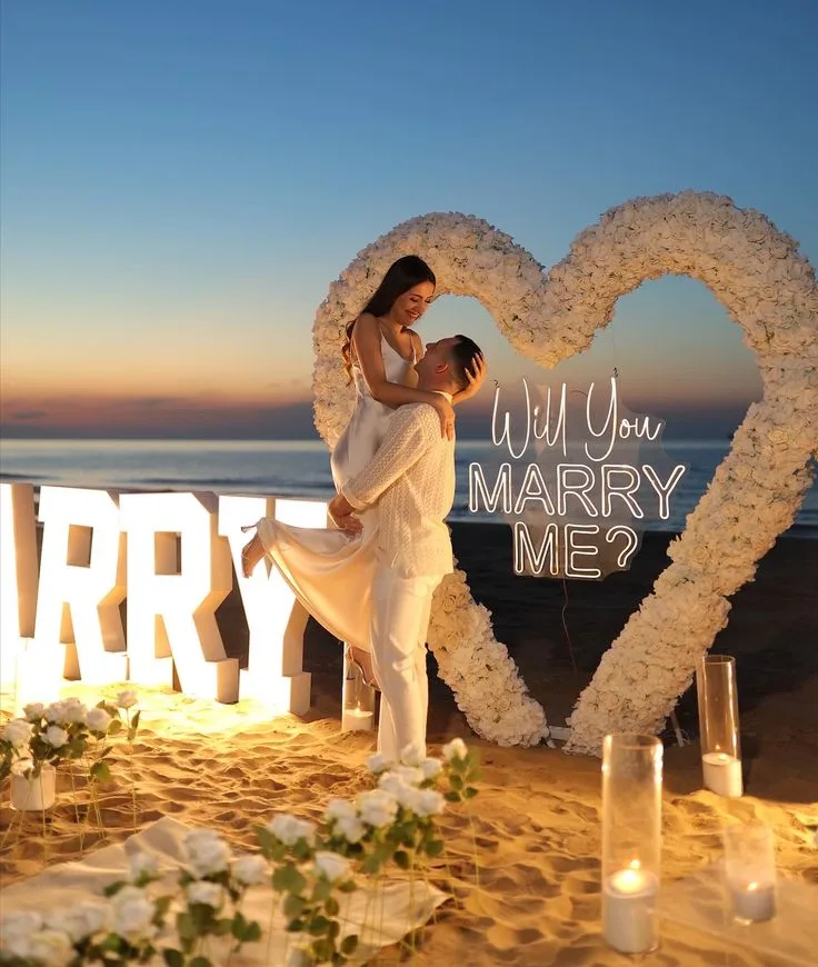 Couple embracing in front of a white floral heart arch and illuminated MARRY ME letters on a Cabo beach at sunset
