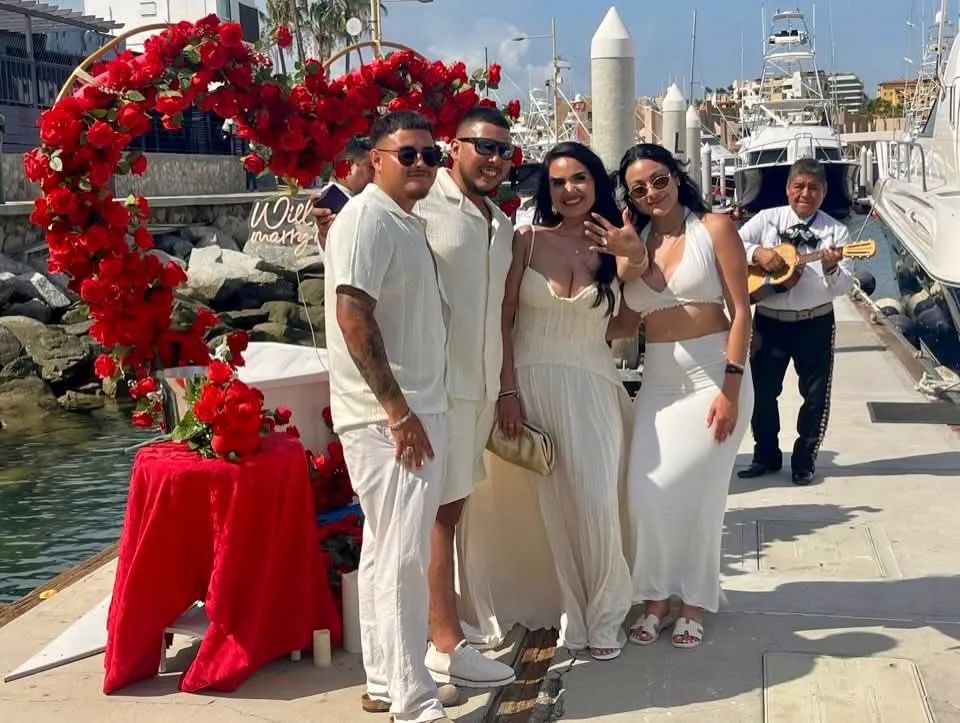Couple and friends celebrating at the marina beside a red rose heart arch with a mariachi player